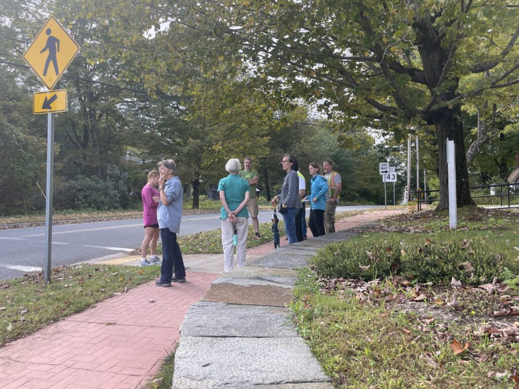A group of people stand at a mid-block crossing with pedestrian signage and discuss conditions. 